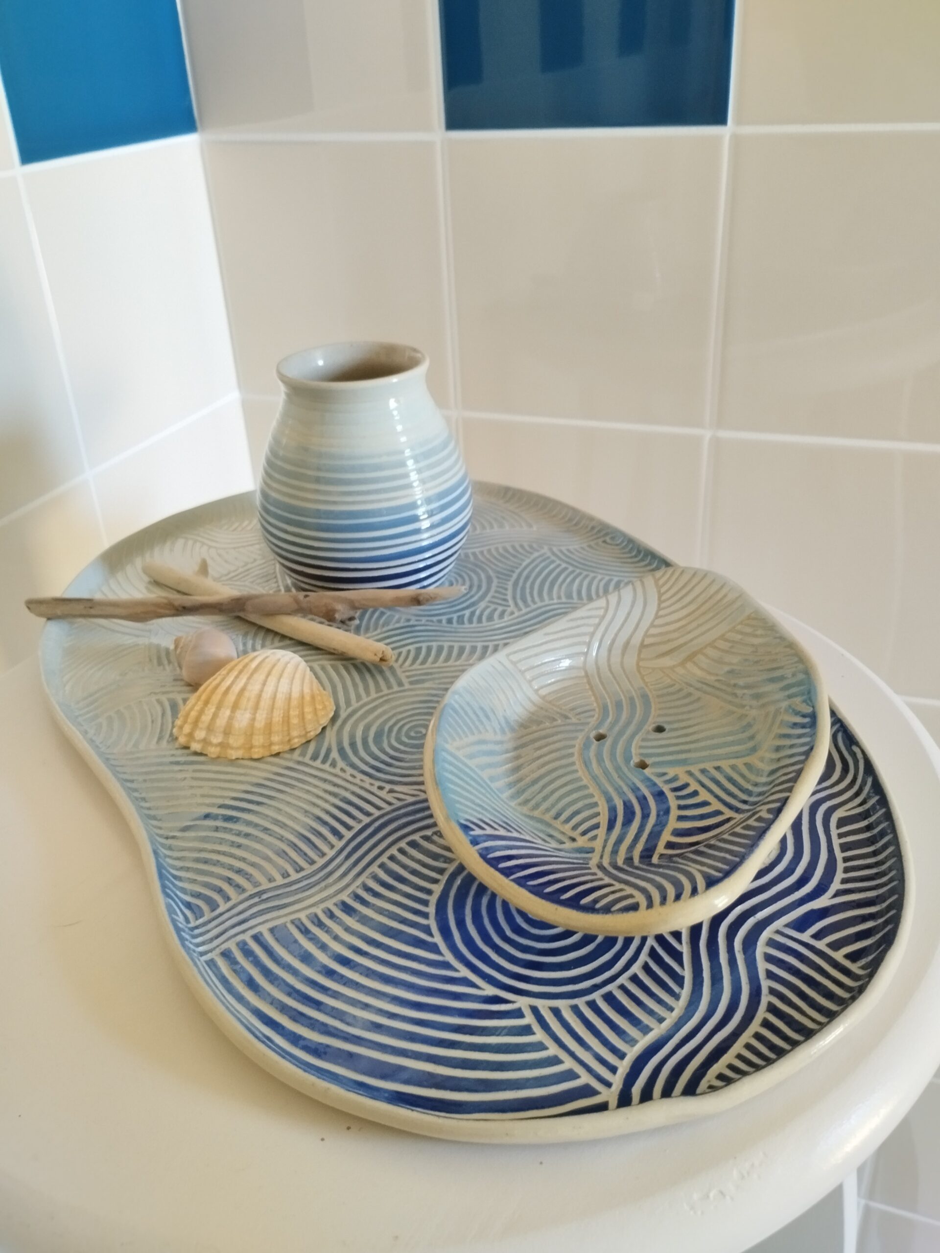 Ensemble de céramique artisanale. Plat, vase et porte-savon peints en dégradé de bleu marine et bleu ciel. Motif de lignes gravées évoquant les mouvements de l'eau. Présentés pour la salle de bain