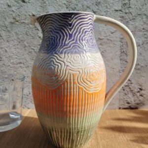 Carafe en céramique artisanale. Fond en dégradé de violet, orange, vert. Motif gravé de fleurs