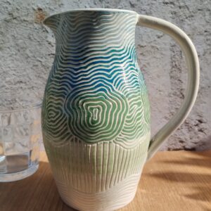 Carafe en céramique artisanale. Fond en dégradé bleu, vert, rose pâle. Motif gravé de silhouettes d'arbres.